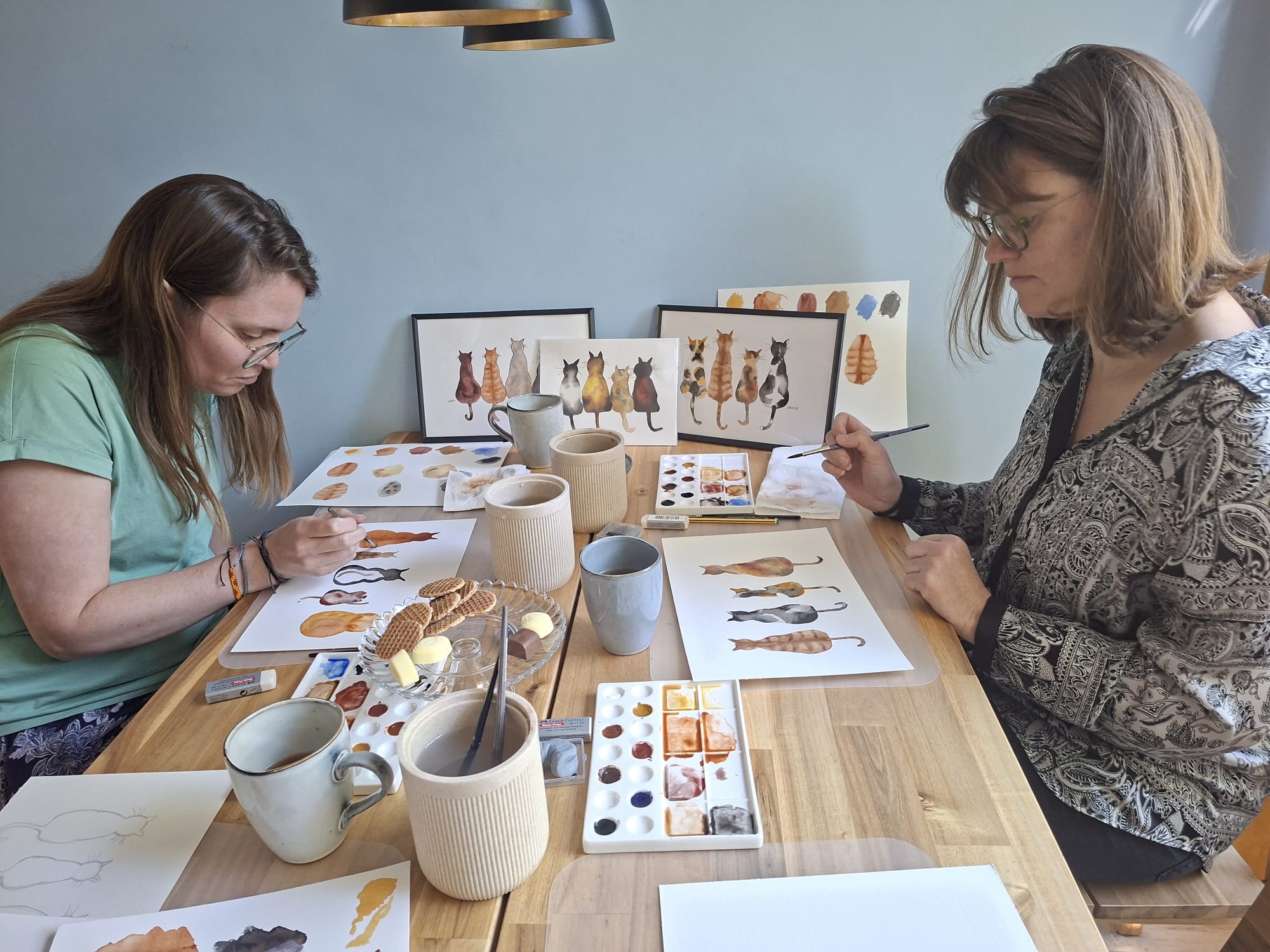 Twee volwassen deelnemers schilderen hun eigen kattenvrienden aan Vera's keukentafel